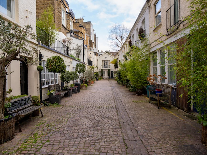 A narrow residential street with cobblestone paving, flanked by white and beige townhouses featuring small balconies with black wrought iron railings and large windows. The street is decorated with potted plants, including small trees and shrubs, placed along the facades of the buildings and on window sills. There are two wooden benches positioned on the pavement, one on each side, and a few plastic containers and additional potted plants near the entrances. In the background, a white building with a central doorway and windows can be seen at the end of the street, suggesting a courtyard or cul-de-sac. The daylight is bright, with a partly cloudy sky overhead, providing natural, diffuse lighting. This setting, as featured by manwithavanwestkensington.co.uk for West Kensington station removals access tips for narrow streets, illustrates a typical city location suitable for home relocation or furniture transport operations, highlighting the tight urban environment that requires careful planning during moving and loading processes.