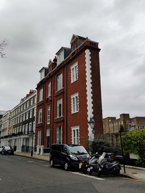 A multi-storey red brick building with white window frames and decorative white accents running vertically along the corner, situated on a street in West Kensington. The street includes parked vehicles, such as a black van and several scooters, with a pavement lined by a low fence and greenery. The overcast sky casts diffuse light over the scene. This location relates to house removals and moving logistics as part of urban furniture transport and packing processes, reflecting the typical environment where [COMPANY_NAME] offers property relocation services, particularly in narrow or challenging street access areas like West Kensington station vicinity.